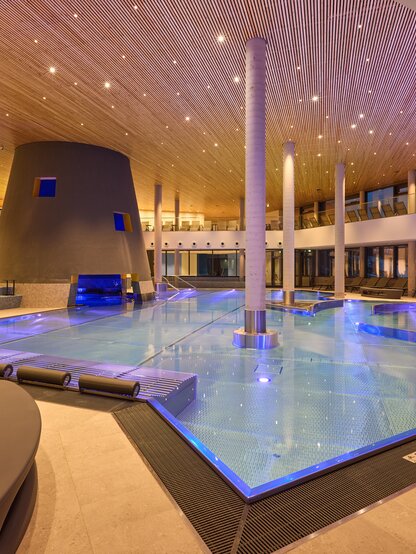 Modern indoor pool with blue lighting, wooden ceiling, and loungers