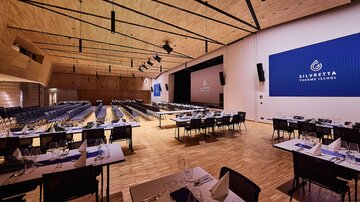 Veranstaltungssaal mit gedeckten Tischen, zwei Leinwänden und Holzdecke, vorbereitet für Event