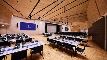Großer Veranstaltungssaal mit gedeckten Tischen, holzverkleidung und Leinwand