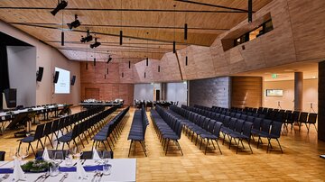 Seitenansicht: großer, moderner Veranstaltungssaal mit Bühne, Leinwand, Reihen schwarzer Stühle und gedeckten Tischen unter Holzdeck