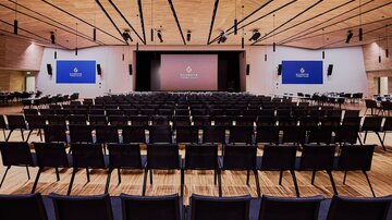 Veranstaltungssaal mit Bühne, Leinwand und Reihen schwarzer Stühle, vorbereitet für Konferenz