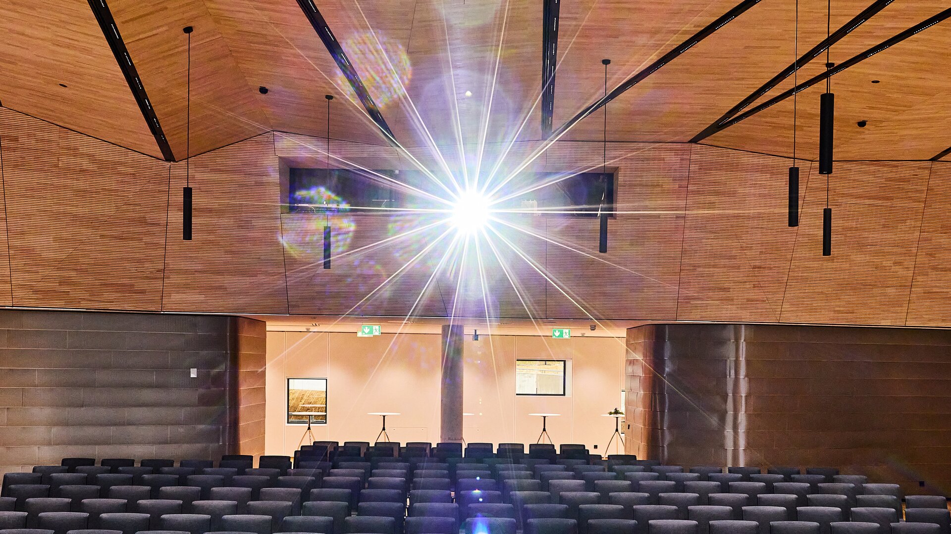 Veranstaltungssaal mit schwarzen Stuhlreihen, Holzdecke und zentralem Lichtstrahl