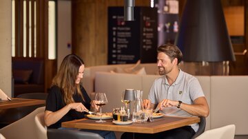 Ein Mann und eine Frau beim Essen in der Grill Lounge, gemütliche Atmosphäre mit stilvoll gedecktem Tisch und warmem Holzdekor.