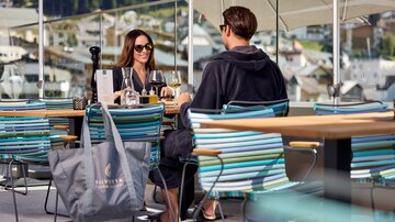 Zwei Personen sitzen im Außenbereich des Restaurants mit Blick auf die Berge, bunte Stühle und sommerliche Atmosphäre.