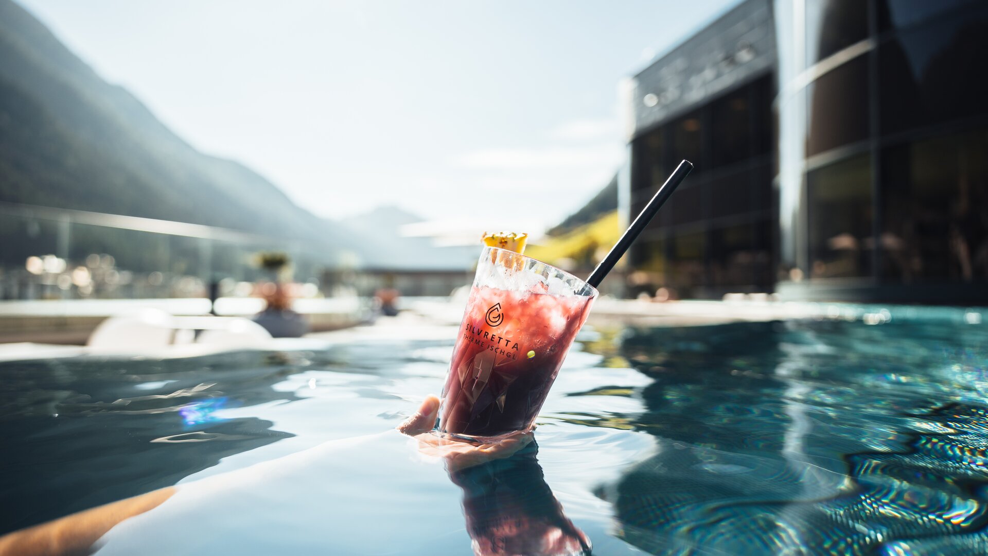 Hand holding red drink in the pool, modern glass facade and mountains in the background