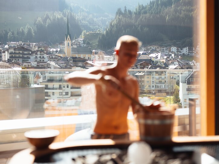 Sauna master with bucket and ladle in the sauna, through a window you can see the valley