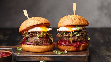Two burgers with fried egg, green topping and red sauce on wooden board